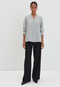 Blouse gris clair à manches longues avec un décolleté en V et une finition texturée subtile, associée à un pantalon noir à jambes larges et des chaussures noires pointues.
