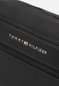 El bolso cruzado de nylon negro presenta una superficie lisa, una cremallera prominente y una banda texturizada con el logo "TOMMY HILFIGER" en plata.