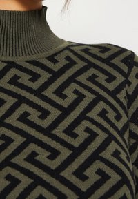 Strickpullover mit hohem Kragen in olivgrün, versehen mit einem auffälligen schwarzen geometrischen Muster. Weiche Textur und figurbetonter Schnitt.