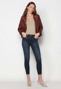 Giacca in pelle bordeaux, top beige senza maniche e jeans skinny blu scuro. Sandali con tacco trasparente completano l'outfit, caratterizzati da un design aderente.