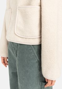 Primer plano de una chaqueta de forro polar color beige claro con grandes bolsillos frontales sobre pantalones verdes desgastados y una mano relajada al lado.