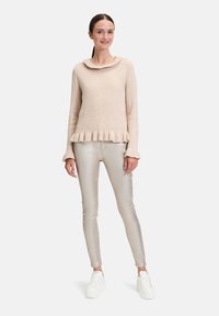 Pull beige côtelé avec un col ondulé et un ourlet évasé. Associé à un pantalon métallique slim et des baskets blanches à plateforme. Le modèle se tient droit.