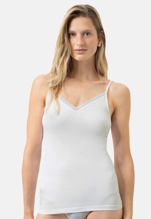 Blonde Frau trägt eine weiße Spitzen-Camisole und passende Unterwäsche, steht vor hellem einfarbigem Hintergrund.