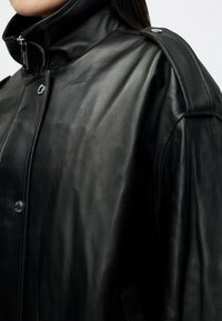 Giacca in pelle nera con colletto alto, hardware argentato e texture sottile, con accenti evidenti sulle spalle e una vestibilità sartoriale.