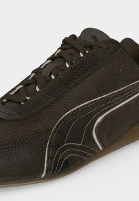 Παντόφλα Puma από σκούρο καφέ δέρμα με υφή, σκούρα κορδόνια, λευκές λεπτομέρειες και καουτσούκ σόλα, εμφανής σε λευκό φόντο.