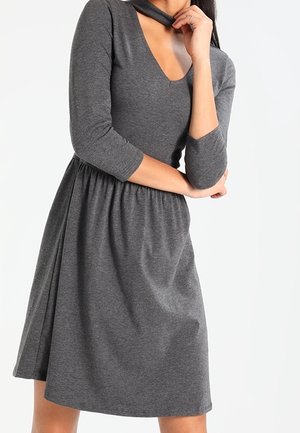 Frau trägt ein graues, knielanges Kleid mit dreiviertellangen Ärmeln und V-Ausschnitt mit Schlitzdetail, posiert mit verschränkten Armen.