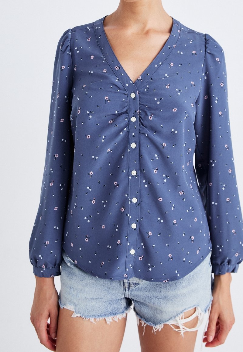 Femme portant un chemisier bleu à manches longues avec de petites impressions florales et un short en denim clair effiloché.