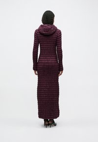 Μακρύ, εφαρμοστό παλτό σε χρώμα δαμάσκηνο, με ποικιλία από επενδύσεις και κουκούλα. Διαθέτει διαρκές σχέδιο από οριζόντιες ραφές και μακριά μανίκια. Φοριούνται μαύρες γόβες.