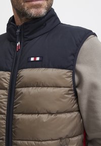 Gepolsterte Weste mit einem schwarzen oberen Abschnitt und einem braunen unteren Abschnitt. Verfügt über einen hohen Kragen, einen kontrastierenden Reißverschluss und ein kleines Logopatch.
