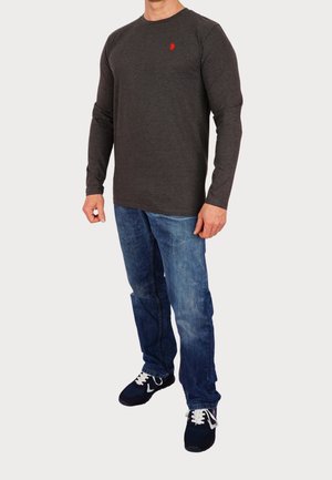 Langarm-Dunkelgraues Shirt mit Rundhalsausschnitt, rotem Logodetail auf der Brust, kombiniert mit hellblauen Jeans und schwarzen Sportschuhen.