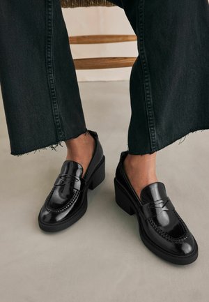 Schwarze lackierte Lederloafers mit einem klobigen Absatz, die über geflochtene Details entlang des Schafts und eine glatte, glänzende Oberfläche verfügen.