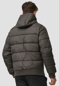 Homme portant une veste à capuche rembourrée gris foncé avec des poignets et un ourlet côtelés, vu de dos sur un fond gris clair uni.