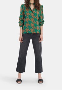 Blusa a motivo houndstooth verde e marrone con scollatura a V profonda, maniche arricciate e vestibilità rilassata. Abbinata a jeans neri a gamba larga e sandali neri.