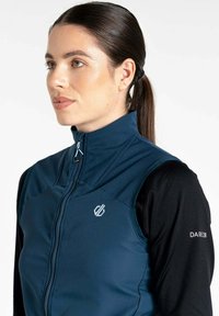 Blaues ärmelloses Jackett mit hochgeschlossenem Kragen, Reißverschluss und Logo auf der Brust; kombiniert mit einem schwarzen Langarmshirt darunter.