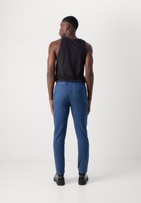 Pantalones azules de tela suave, con corte ajustado, frente plano y dos bolsillos traseros. Combinados con una camiseta sin mangas negra y zapatos negros.