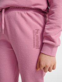 Primo piano della mano di una persona che riposa lungo il fianco, indossando pantaloni della tuta rosa hummel e manica di felpa coordinata.