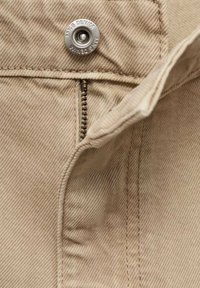 Beige denim fabric with visible texture, featuring a metal button closure and zipper. Seam details along the edge are prominent.