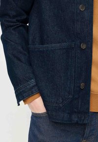 Giacca in denim blu scuro con tasca sul petto, chiusura con bottoni e cuciture a contrasto. Lo strato interno è una felpa marrone.