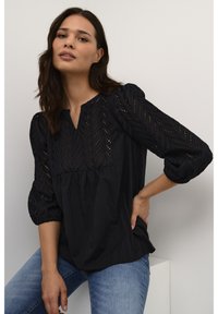 Svart blus med ¾-lång puffärm, prydd med ett chevronhålmönster och en avslappnad passform, kombinerad med ljusa denimjeans.