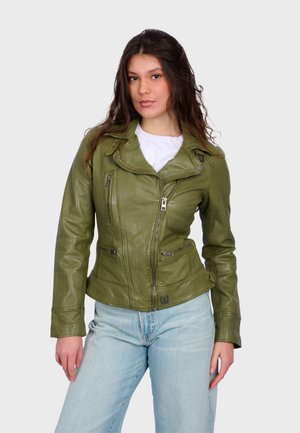 Nuori nainen, jolla on pitkät hiukset, pukeutuneena oliivinvihreään nahkatakkiin valkoisen paidan ja vaaleansinisien farkkujen päällä, seisoo yksinkertaisen taustan edessä.