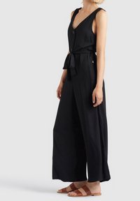 Black sleeveless jumpsuit with a deep V-neck, front buttons, and a waist tie. Features wide legs and side pockets; made of lightweight fabric.