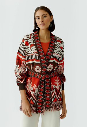 Vrouw draagt een rood, bruin en wit gebreid vest met een ceintuur, een franjeszoom en bloemen- en geometrische patronen, gecombineerd met een witte broek.