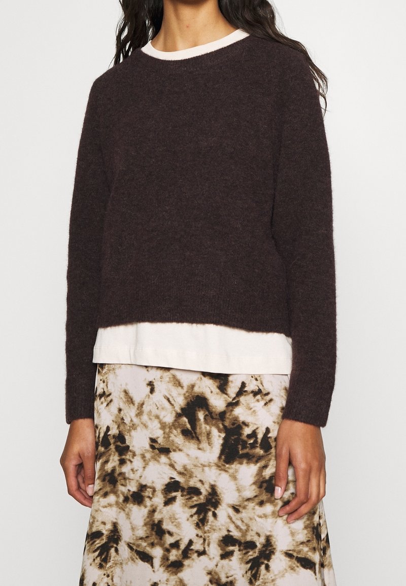 Eine Frau trägt einen dunkelbraunen, flauschigen Pullover über einem weißen Hemd, kombiniert mit einem beige-schwarz gemusterten Rock mit abstraktem Design.