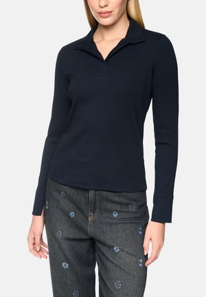 Woman wearing a long-sleeve navy shirt with a collar and dark jeans embroidered with small blue flowers, standing against a plain background.