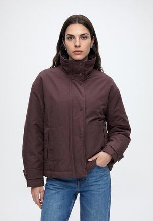 Kvinde med langt mørkt hår iført en brun jakke med høj krave og blå jeans, står med den ene hånd i lommen mod en ensfarvet baggrund.