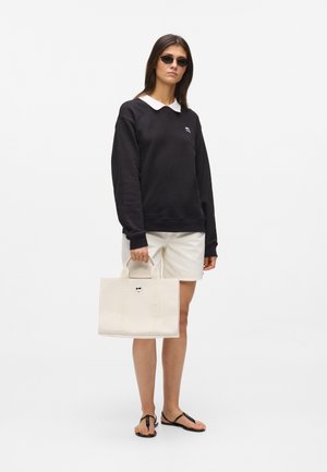 Ženska, ki nosi črno dolg rokav s belim ovratnikom, bele kratke hlače, črna sončna očala, črne sandale in drži bež torbo z ročaji.