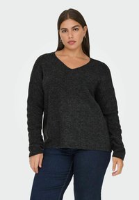 Temno siv pulover iz pletenine s V-izrezom, dolgimi rokavi in sproščenim krojem, v kombinaciji s temno modrimi jeans hlačami. Teksturirana tkanina in sproščen dizajn.