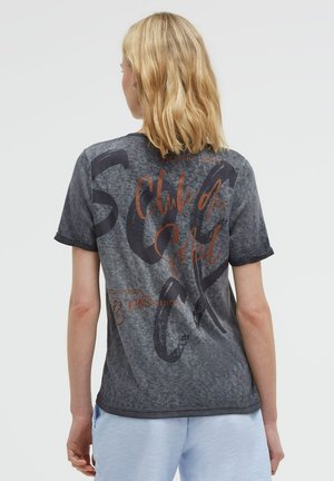 Blonde Person trägt ein ausgewaschenes schwarzes Grafik-T-Shirt mit Text und hellblaue Shorts, steht mit dem Rücken zu einem schlichten Hintergrund.