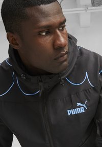 Czarna kurtka z zamkiem błyskawicznym i niebieskimi akcentami, z logo PUMA na klatce piersiowej, z kapturem i gładką, lekką teksturą.