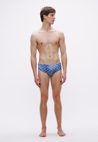 Bañador slip de hombre en tela azul con un sutil patrón geométrico. El diseño presenta una cintura baja y un ajuste ceñido.