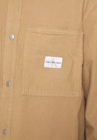 Camicia di cotone beige con bottoni a pressione, dotata di una tasca sul petto con un'etichetta bianca che riporta la scritta "Calvin Klein Jeans." Tessuto liscio, design classico.