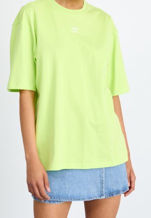 Persoon draagt een losse lichtgroene Adidas T-shirt en een blauwe denim minirok, staand tegen een effen witte achtergrond.