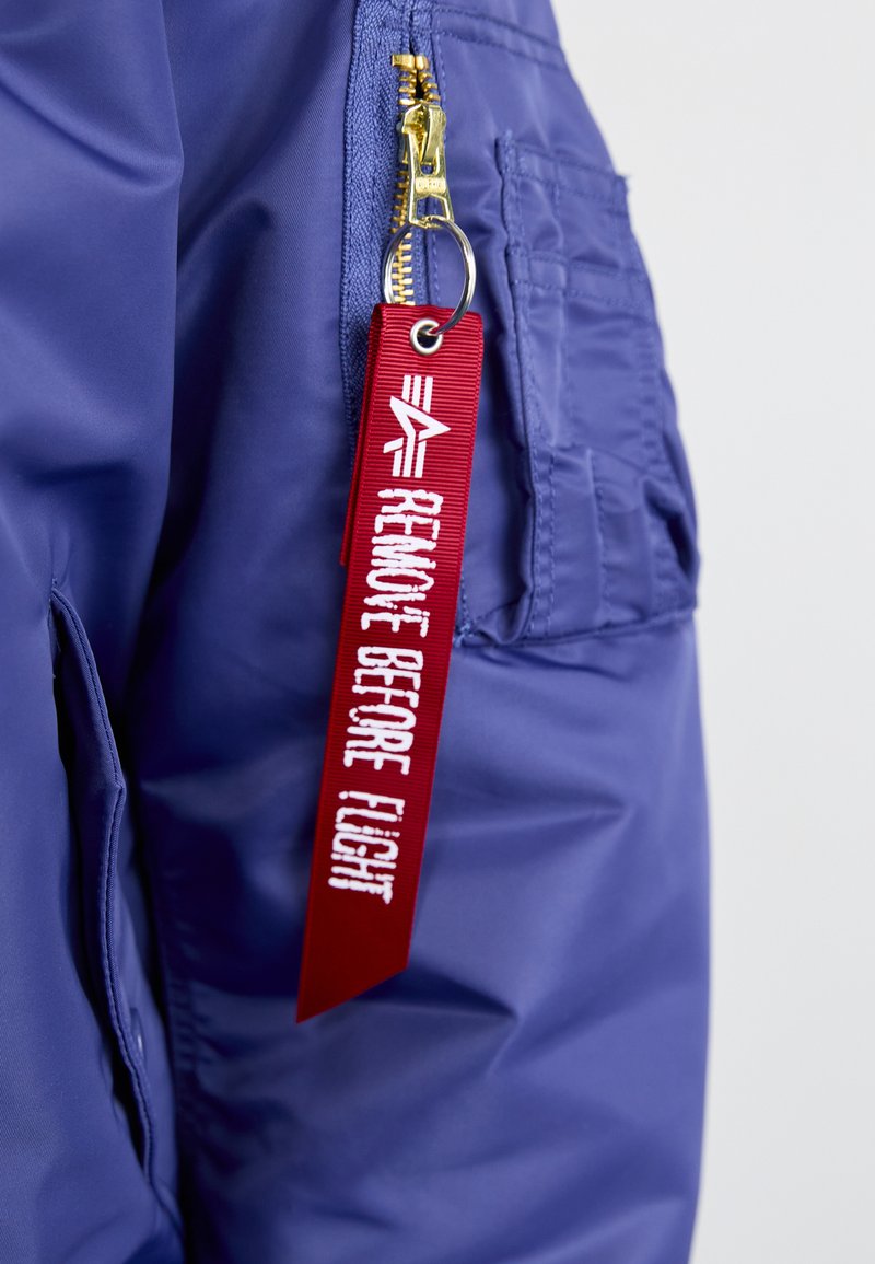 Giacca bomber di colore blu navy con texture lucida, dotata di una cerniera dorata e di un'etichetta di tessuto rossa che recita "RIMUOVERE PRIMA DEL VOLO".