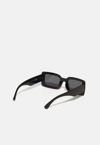 Lunettes de soleil noires avec des montures rectangulaires, des verres gris et une texture en plastique lisse. Présentent des branches allongées et courbées ainsi qu'une subtile marque à l'intérieur.
