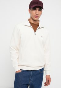 Pullover di pile bianco con colletto a zip quarti, dotato di un piccolo logo sul petto e abbinato a jeans in denim blu. Incluso un cappellino bordeaux.