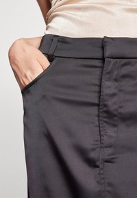 Närbild av en hand som vilar i framfickan på svarta skräddarsydda byxor som bärs med en ljusbeige topp.