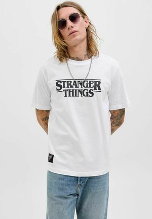 Bílý tričko s krátkým rukávem s nápisem "STRANGER THINGS" tištěným černě. Má kulatý výstřih a malou značkovou štítek na boku.