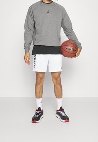 Felpa grigia a maniche lunghe con logo nero, sottocamicia nera, pantaloni corti da basket bianchi in rete con brand laterale, sneaker nere e rosse, e pallone da basket.