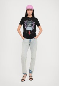 Black t-shirt featuring a multicolored graphic of a birdcage with flowers, paired with light-wash jeans and a pink cap. Black heeled sandals.