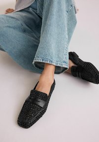 Mocasines negros trenzados con punta cuadrada, que presentan una tira de cuero negro plano en la parte superior. Llevados con unos vaqueros de pierna ancha color azul claro.