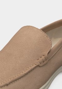 Ανοιχτό καφέ σουέντ παπούτσι χωρίς κορδόνια με ορατές ραφές και λευκή σόλα, εμφανιζόμενο σε κοντινή λεπτομέρεια.