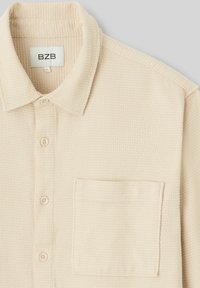 Chemise boutonnée beige clair avec un col, tissu texturé, petite poche plaquée et boutons visibles sur le devant.
