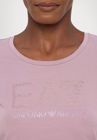 Maglietta rosa chiaro con scollo rotondo e logo di strass scintillanti "EA7 Emporio Armani" al centro. Tessuto morbido.