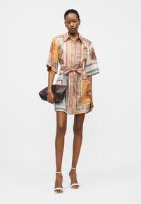 Γυναίκα που φοράει πουκάμισο-φόρεμα με μοτίβο και ζώνη, καθώς και λευκές γόβες, κρατώντας μια μεγάλη σκούρα καφέ τσάντα φάκελο μπροστά σε απλό φόντο.