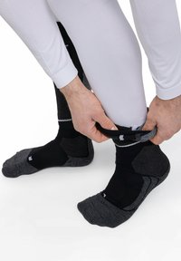 Personne en leggings blancs tirant des chaussettes de sport noires et grises sur les deux pieds sur fond blanc.