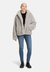 Nepbont jacket in lichtgrijs, met een brede kraag, lange mouwen en zijzakken. Gecombineerd met blauwe jeans en zwarte laarzen.